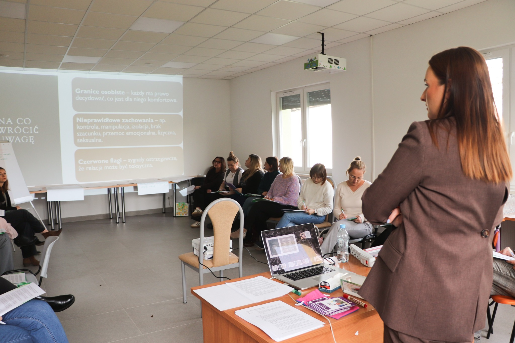 „Prowadząca szkolenie stoi przy stole z laptopem, zwrócona w stronę grupy uczestników siedzących w półokręgu. Na ścianie wyświetlona jest prezentacja dotycząca granic osobistych i nieprawidłowych zachowań.”