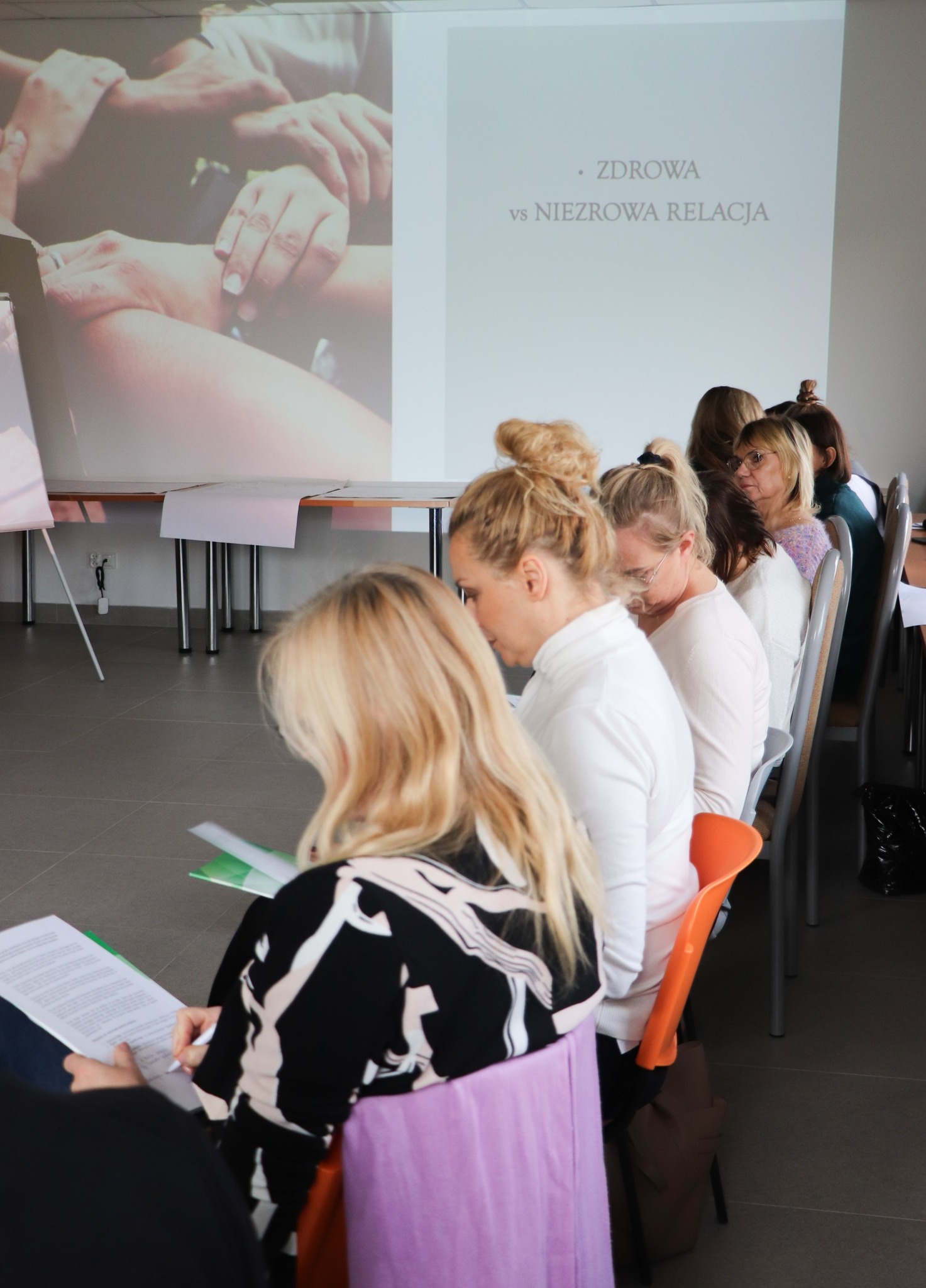 Uczestniczki szkolenia siedzą w rzędzie i robią notatki, a na ścianie przed nimi wyświetlona jest prezentacja dotycząca zdrowej i niezdrowej relacji.