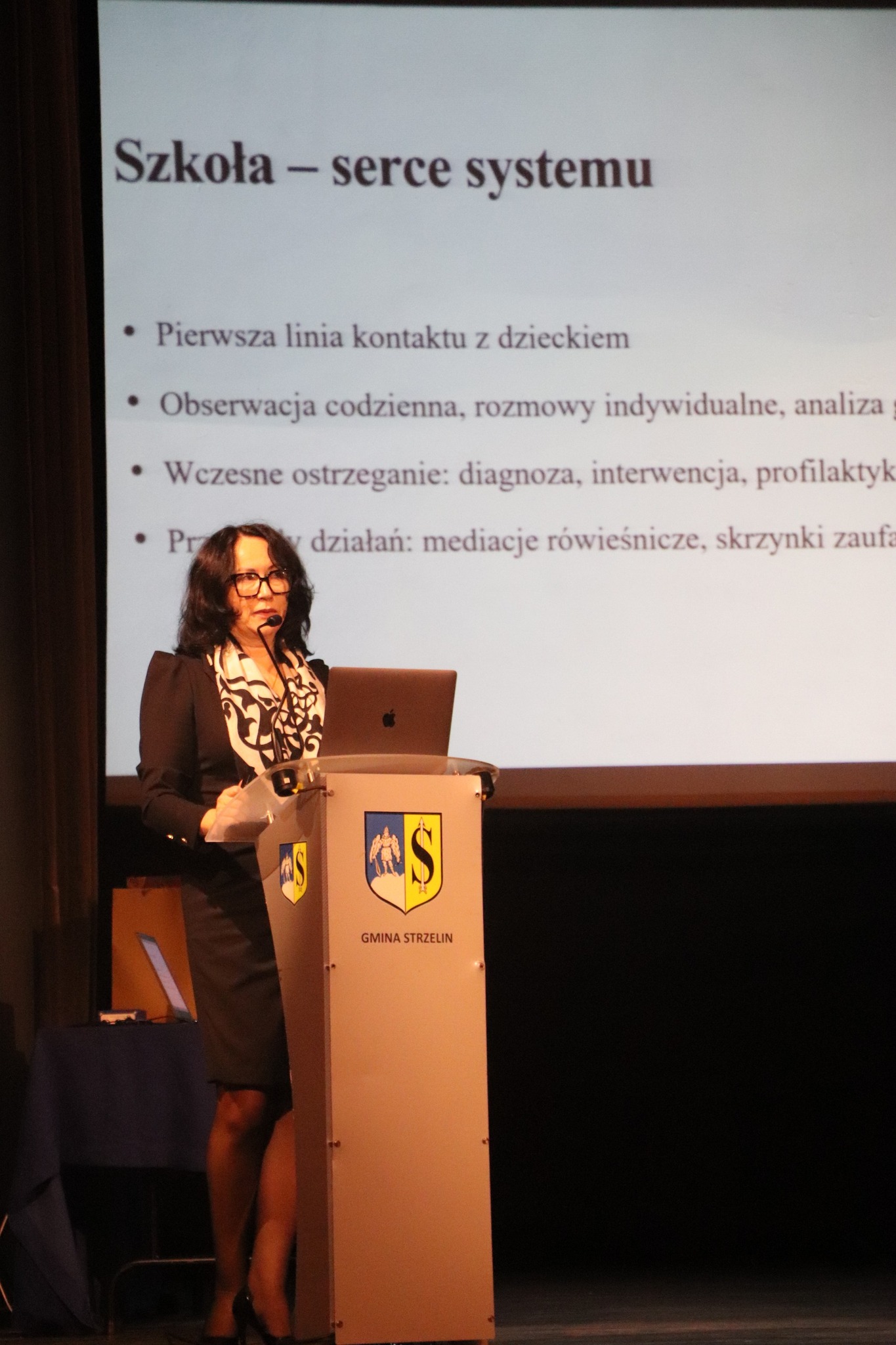 Kobieta prowadzi prezentację na konferencji, stojąc przy mównicy przed publicznością.