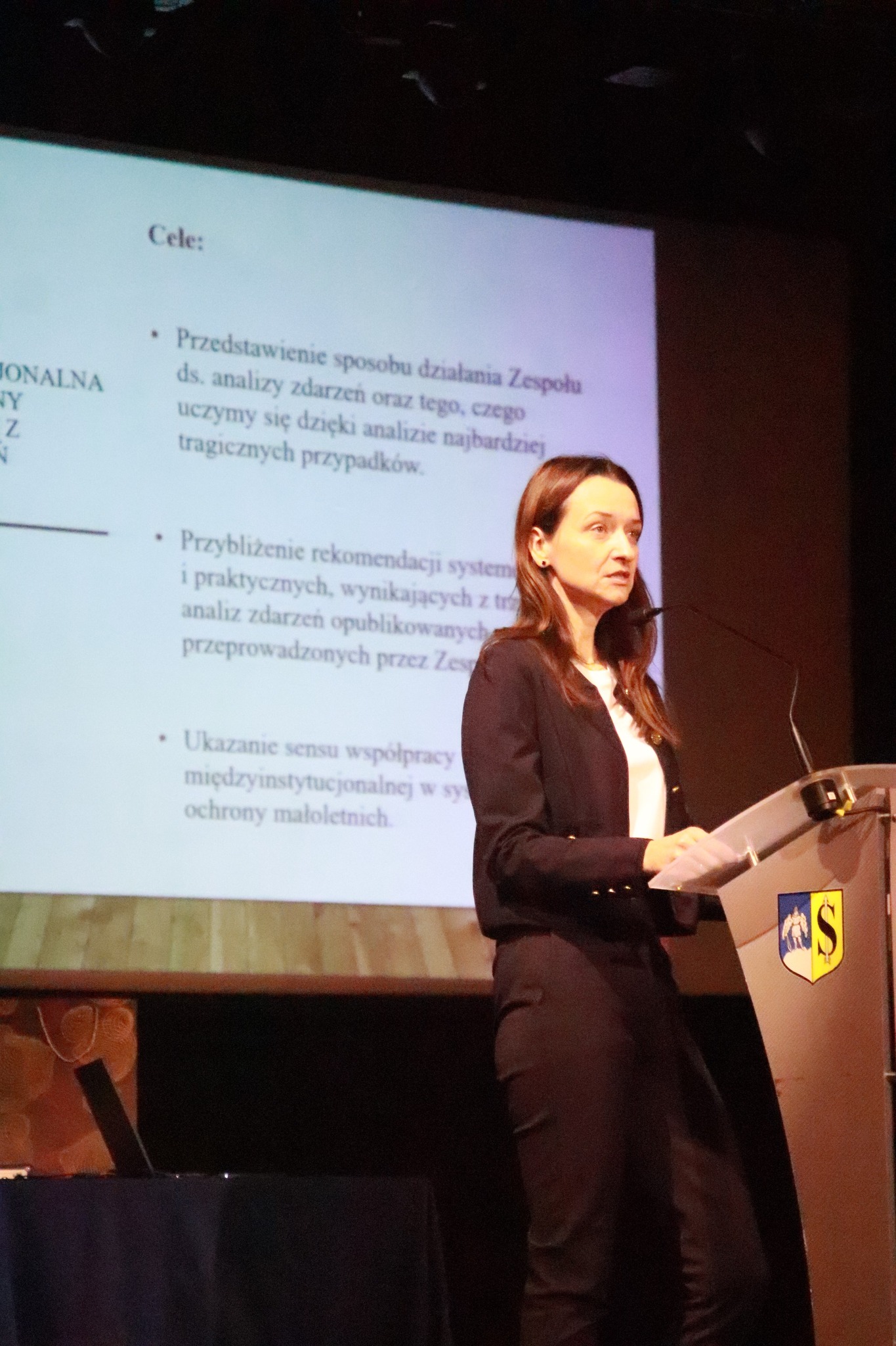 Kobieta prowadzi prezentację na konferencji, stojąc przy mównicy i zwracając się do publiczności.