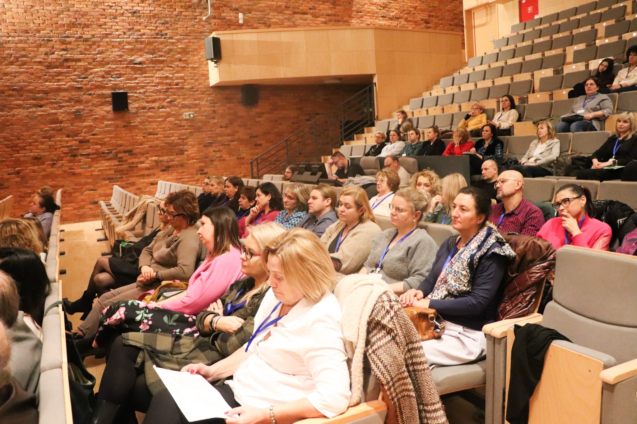 Duża grupa ludzi siedzi w sali wykładowej, zwrócona w stronę prelegenta na scenie.