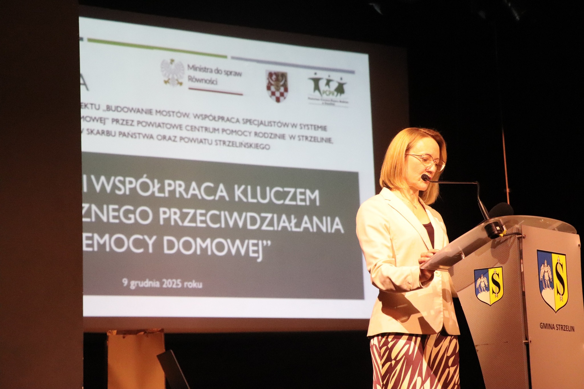 Kobieta wygłasza przemówienie na konferencji, stojąc przy mównicy przed słuchaczami.