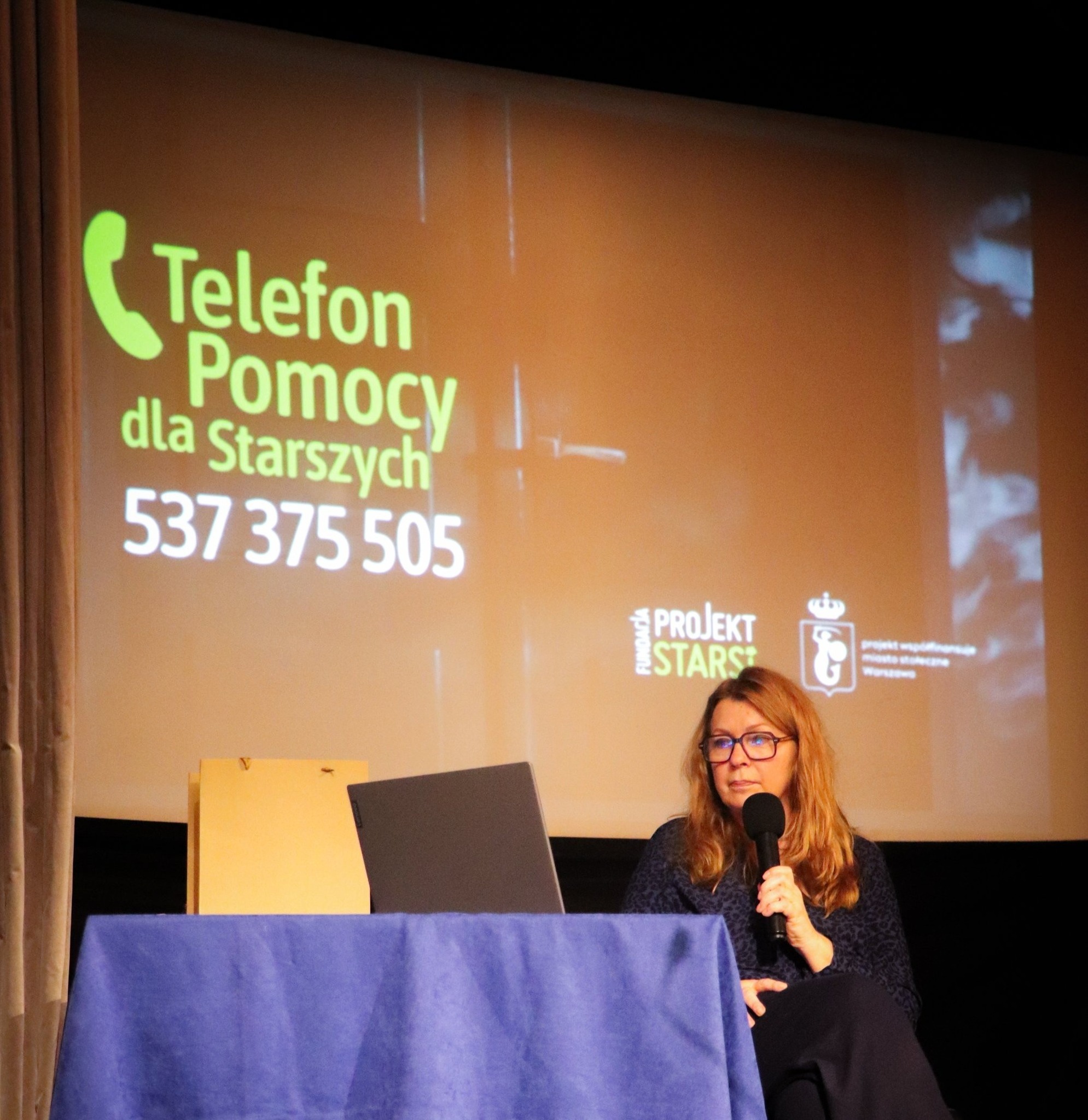 Kobieta przy stole z laptopem i mikrofonem, angażująca się w prezentację.