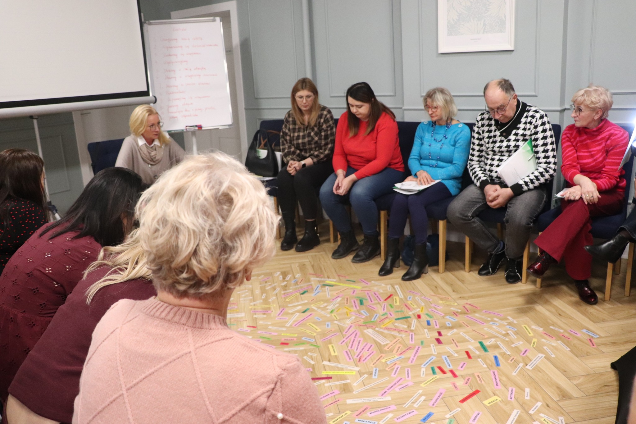 Zespół ludzi siedzących na krzesłach, z widocznymi, różnokolorowymi karteczkami w różnych kolorach porozrzucanymi na podłodze, prowadzi rozmowę.