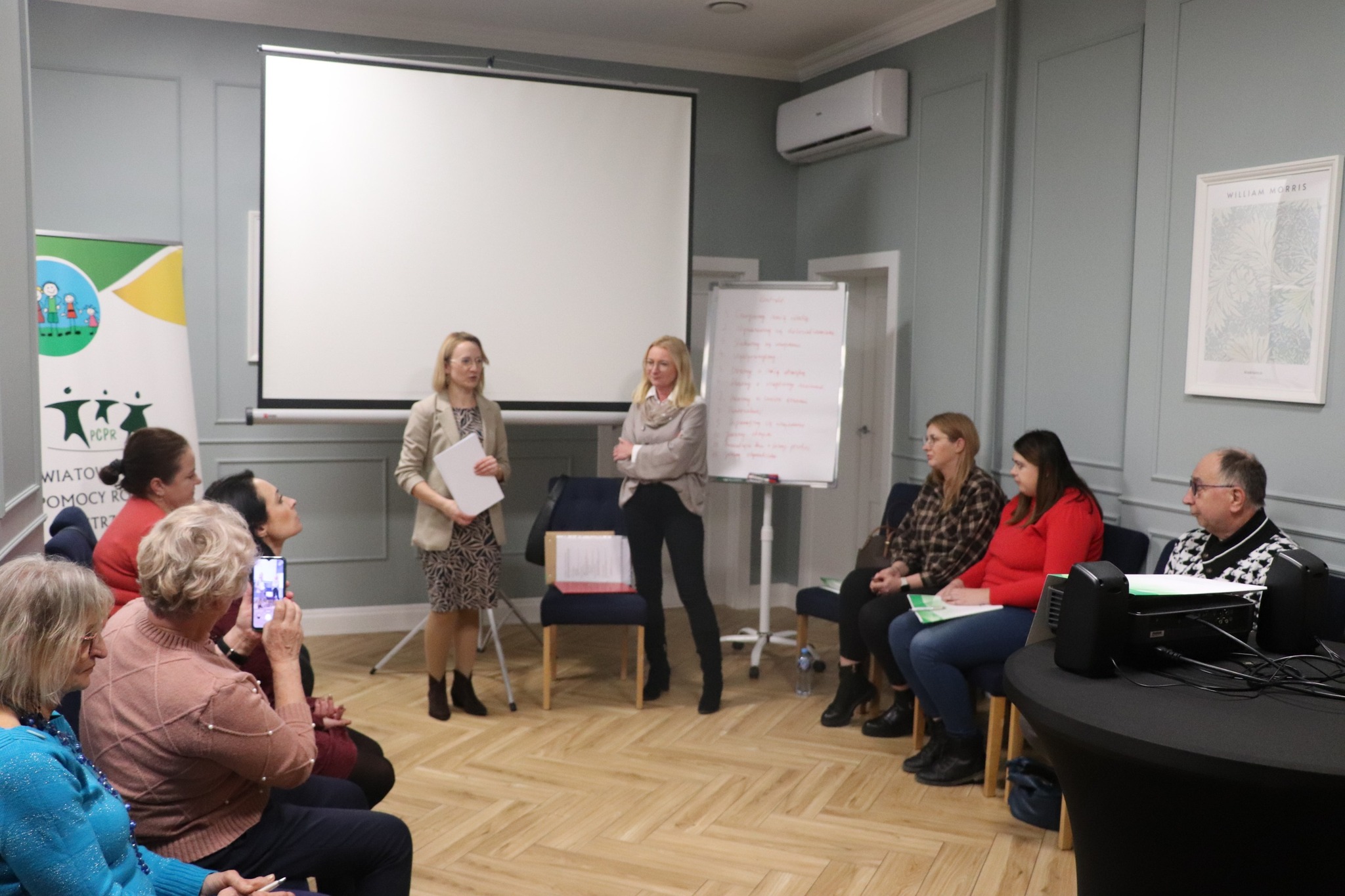 Ludzie zgromadzeni w pomieszczeniu, dwie osoby prowadzą prezentację.