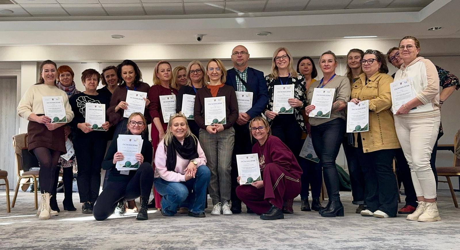 Ludzie w sali trzymają certyfikaty, ciesząc się z sukcesów i wspólnego osiągnięcia.