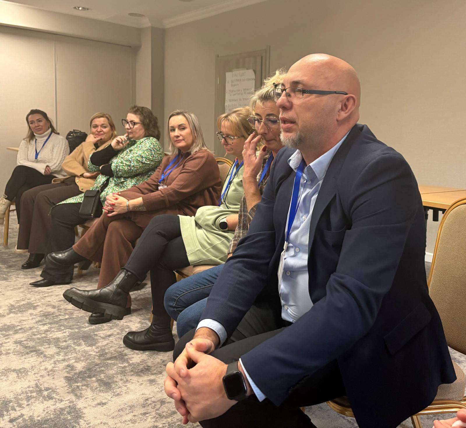 Mężczyzna siedzący w krześle z rękami na kolanach, w tle widoczni słuchacze szkolenia.