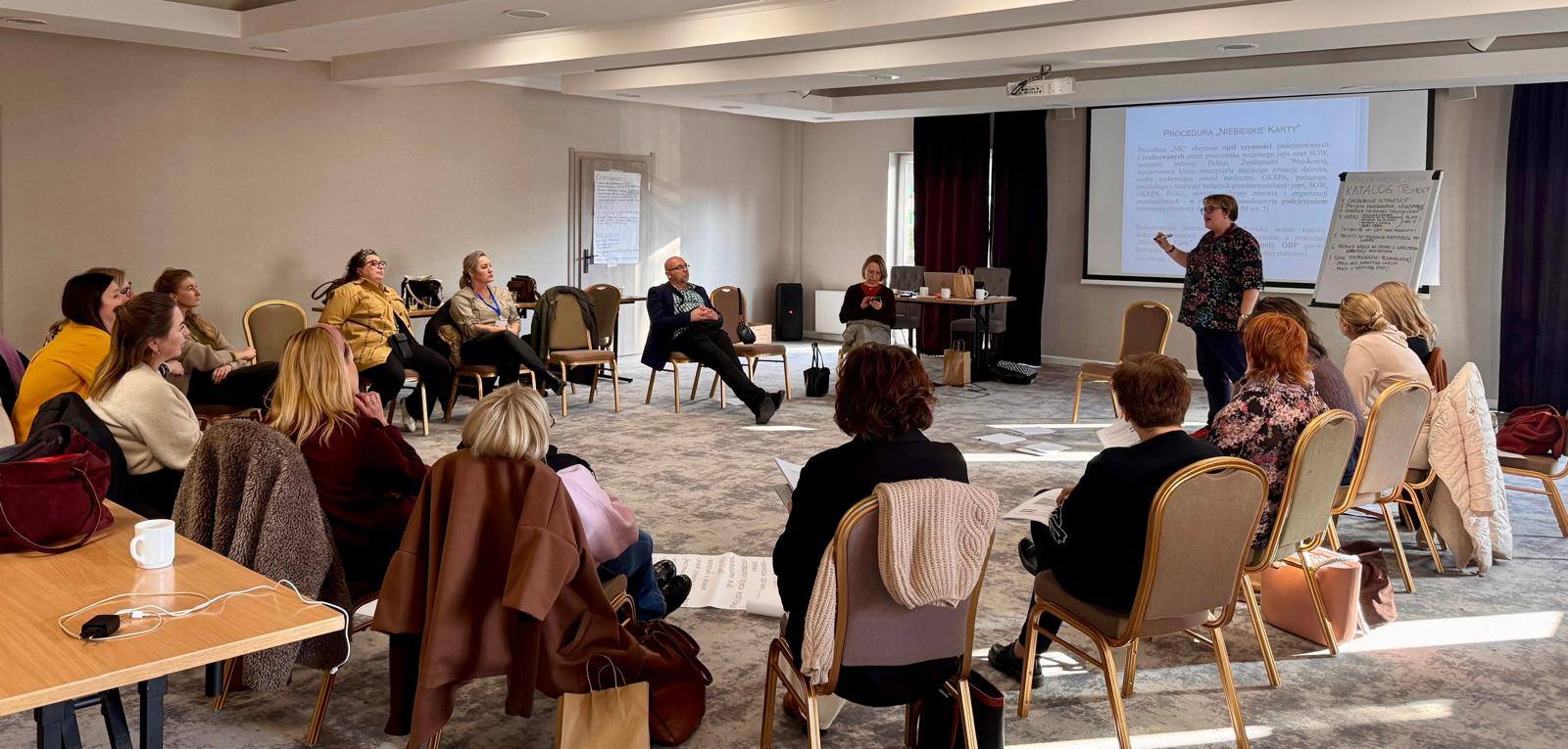 Zespół ludzi siedzących na krzesłach w pokoju, uczestniczący w spotkaniu.