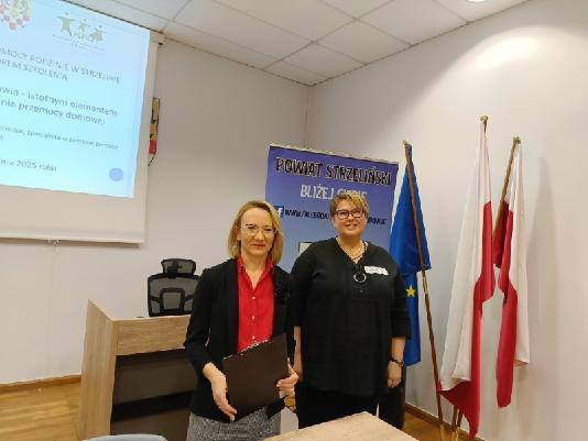 Dwie kobiety stojące przed stołem, w tle widoczna prezentacja wyświetlana na ekranie