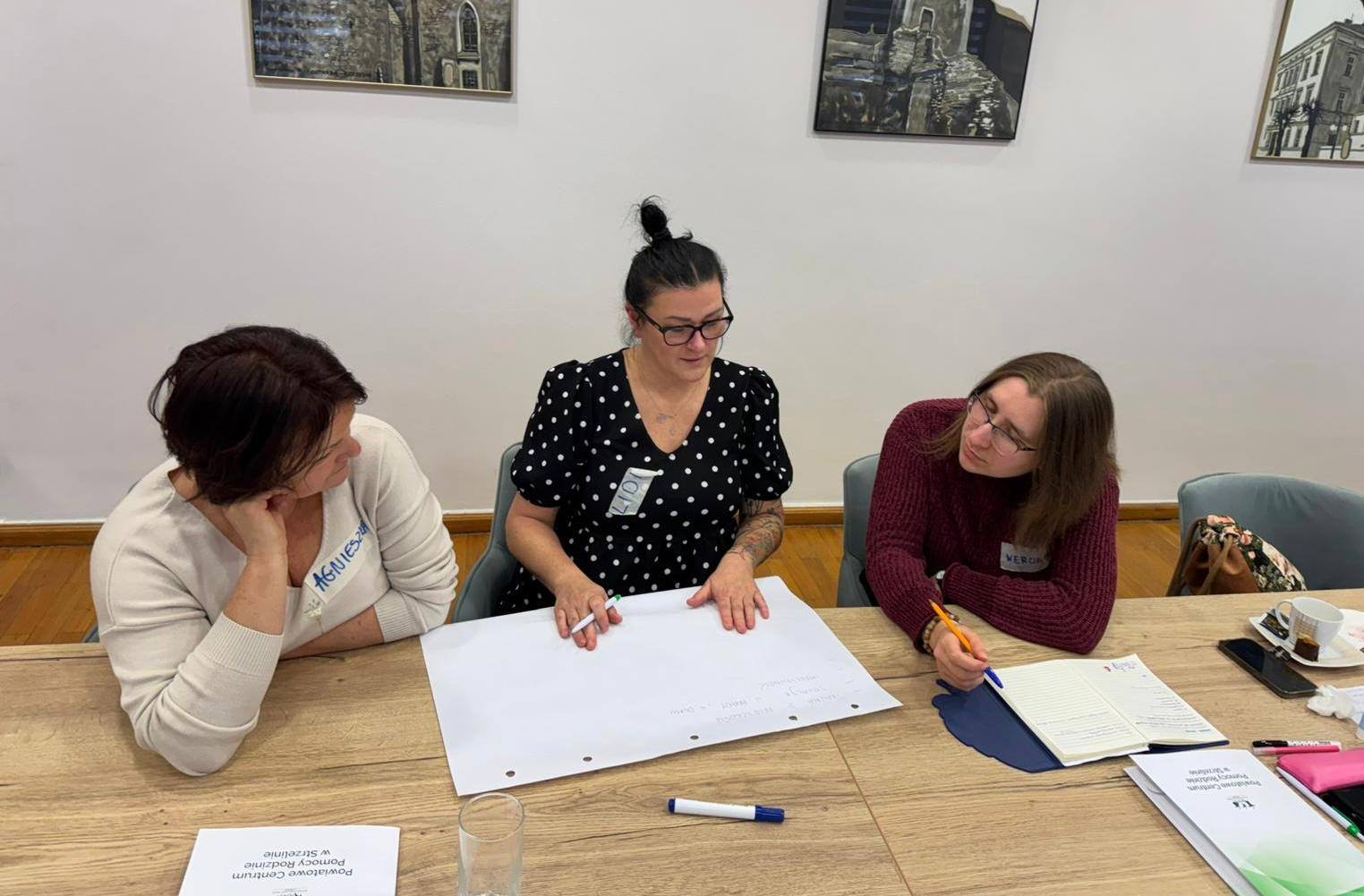 Trzy kobiety przy stole z dokumentami i długopisami, zaangażowane w rozmowę.