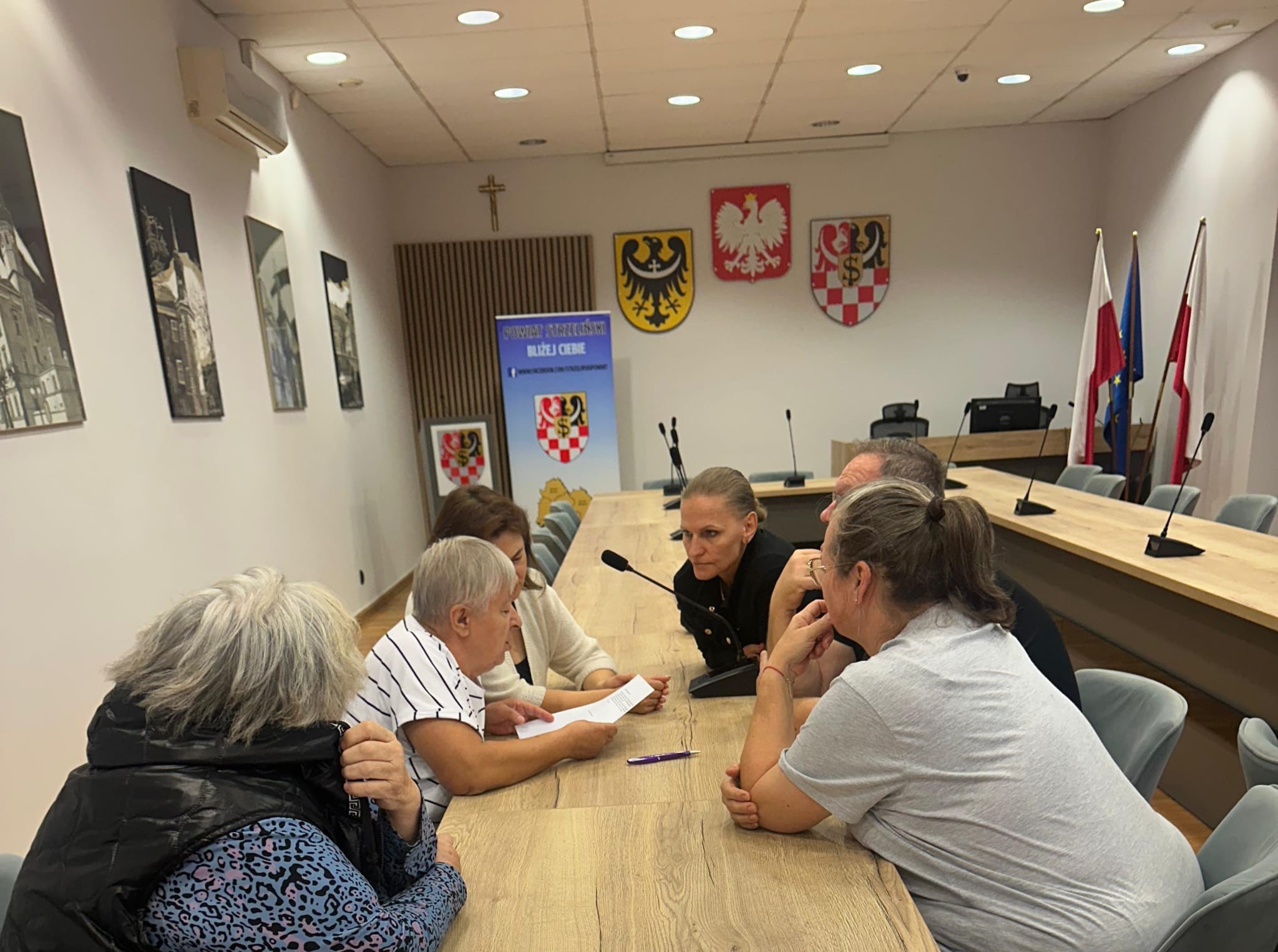 Grupa ludzi siedzi przy stole w sali konferencyjnej podczas spotkania.