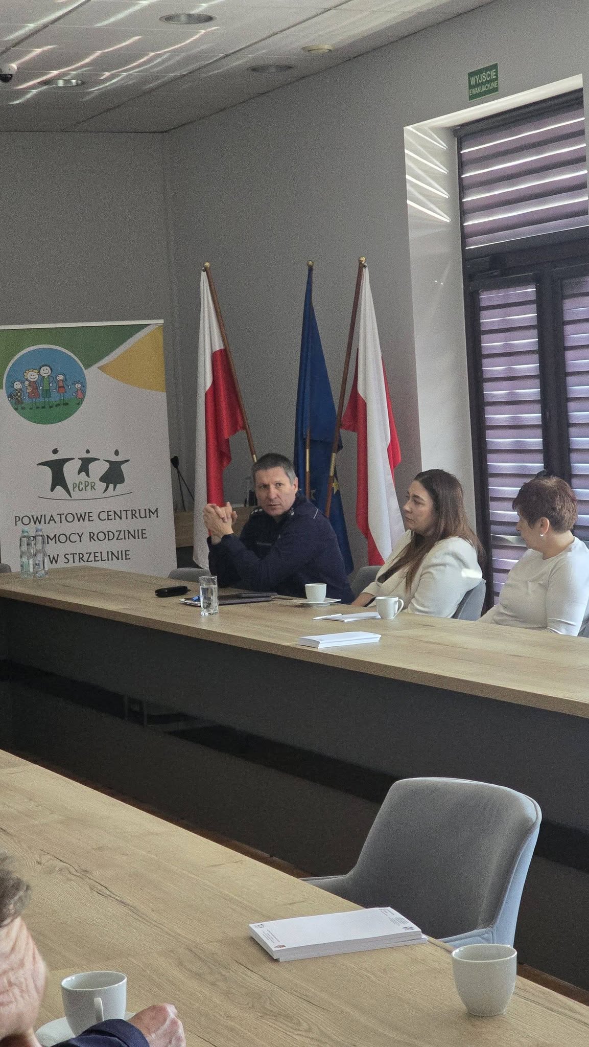 Trzy osoby siedzące przy stole. W tle widoczne są flagi Polski i Unii Europejskiej