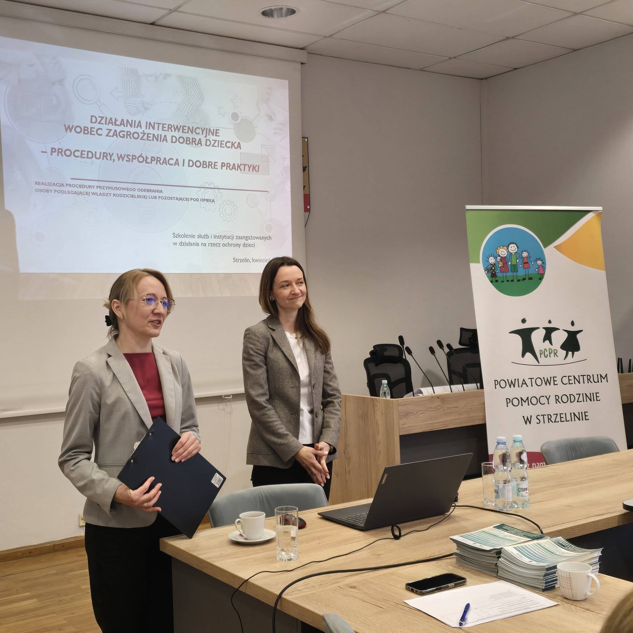 Dwie osoby stoją na tle ekranu, na którym wyświetlana jest prezentacja