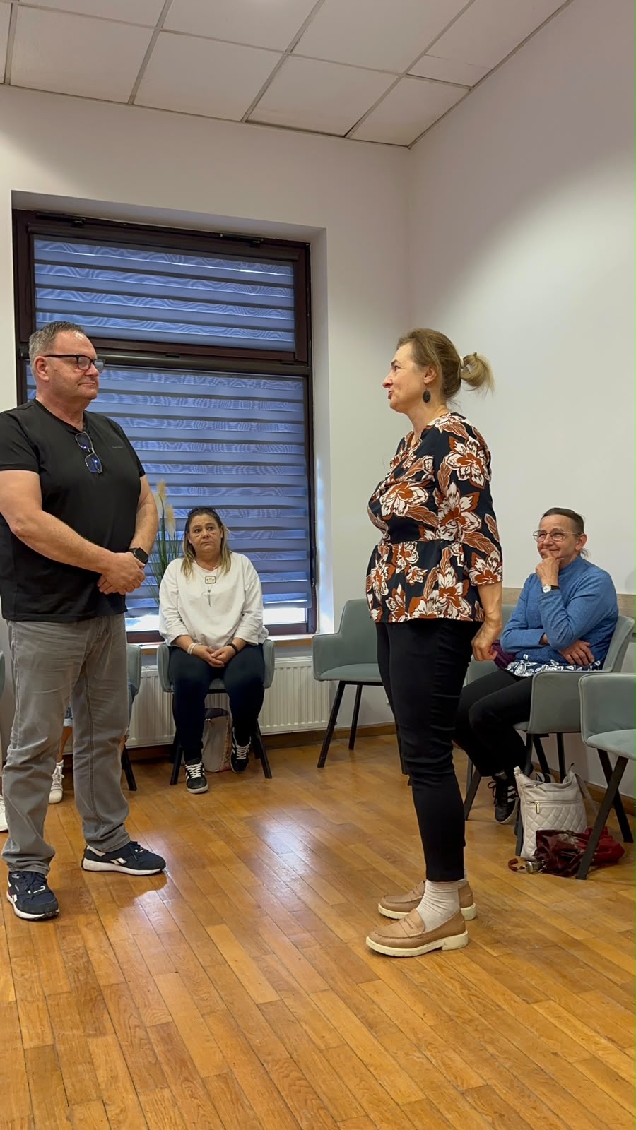 Mężczyzna i kobieta stoją przed grupą ludzi, prowadząc dyskusję lub prezentację.